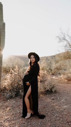 a pregnant woman wearing a long black dress and hat standing in front of a cactus