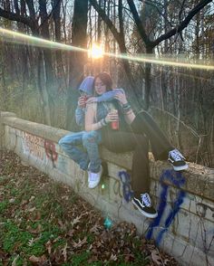 two people sitting on a stone wall with graffiti and trees in the background, one person is hugging the other