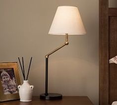 a lamp on a table next to a bed with a picture and an empty vase