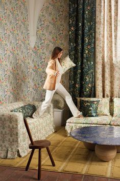 a woman standing in front of a living room with floral wallpaper and patterned curtains