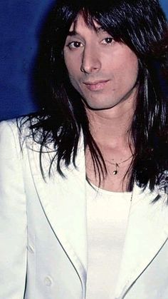 a man with long hair wearing a white blazer and posing in front of a blue background