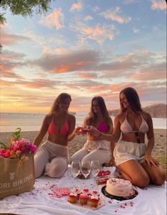 three women in bikinis sitting on a blanket at the beach with cupcakes