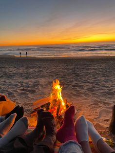 two people are sitting on the beach by a campfire at sunset with their feet up