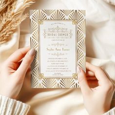 a person holding up a wedding card on top of a white sheet with gold trim