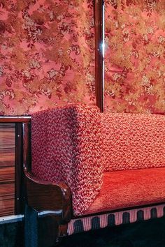 a red couch sitting in front of a wall covered in floral wallpaper and wooden furniture