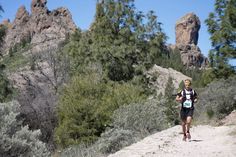 a man running down a dirt road in front of some rocks and trees on a sunny day