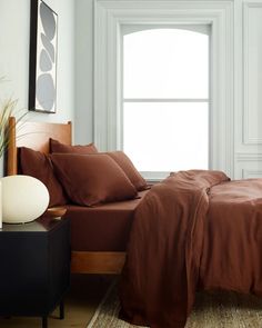 a bed with brown sheets and pillows next to a large window in a white room