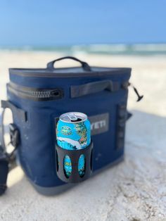 a blue cooler bag sitting on top of a sandy beach next to the ocean with a can of beer in it