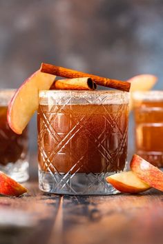 two glasses filled with apple cider on top of a wooden table next to sliced apples