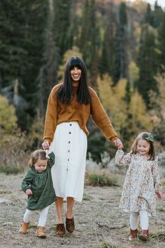 a woman holding hands with two small children in the grass and trees behind her, both wearing brown sweaters