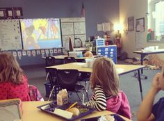 children sitting at desks in a classroom watching cartoons on the television screen and eating lunch