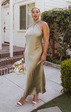 a woman standing on the sidewalk in front of a white house wearing a tan dress