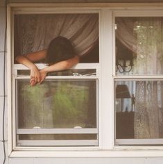 a woman is looking out the window with her head hanging over the curtain and hands on the windowsill