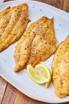 three fish fillets on a white plate with lemon wedges