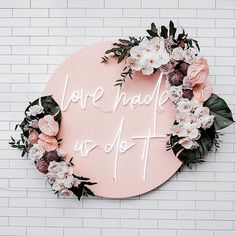 a pink sign that says love makes us do it with flowers around it and greenery