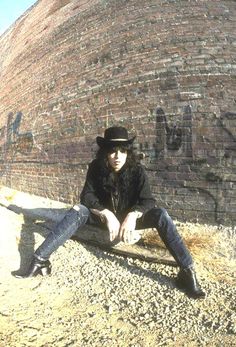 a man sitting on top of a wooden bench in front of a brick wall with graffiti