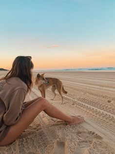 a woman sitting in the sand with a dog on the beach behind her at sunset