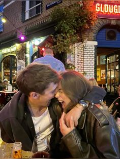 a man and woman sitting at a table in front of a restaurant with their arms around each other