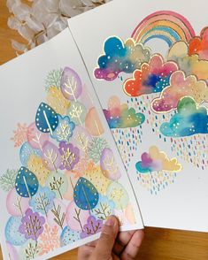 two handmade greeting cards with watercolor clouds and rainbows on them, one being held in front of the other