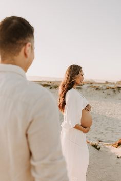 a pregnant woman standing on top of a beach next to a man in a white shirt