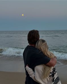 a man and woman hug on the beach as the moon rises over the ocean behind them