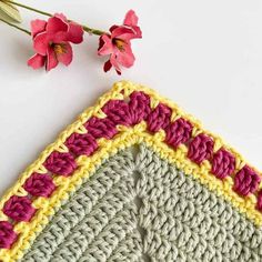 a crocheted square with pink and yellow trim next to red flowers on a white surface