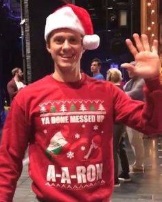 a man in a santa hat waves to the camera while wearing a red sweater and black pants