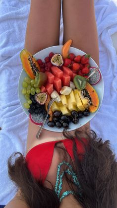 a woman laying in bed with a plate of fruit on her stomach and the bottom half of her body