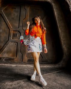 a woman in an orange shirt and white skirt posing for the camera with her hand up