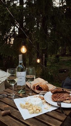 an outdoor picnic table with wine, cheese and crackers on it in the evening