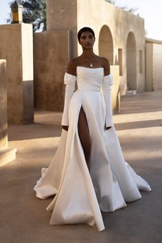 a woman in a white dress with long sleeves and a slited skirt is posing for the camera