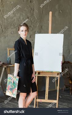 a woman standing in front of an easel holding a paintbrush and looking at the camera