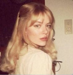 a woman with long blonde hair standing in front of a wall and looking at the camera