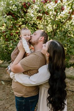 a man and woman holding a baby in front of an apple tree with apples on it