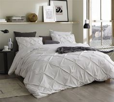 a bed with white comforter and pillows in a room next to a window on a wooden floor