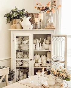 a white china cabinet filled with dishes and vases next to a table topped with flowers