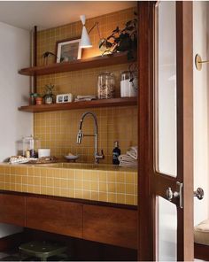 a kitchen with yellow tiled walls and wooden shelves above the sink, along with potted plants