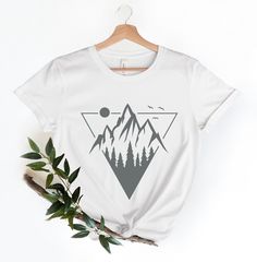 a white t - shirt with mountains and trees on the front is hanging from a wooden hanger