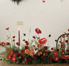 an arrangement of red flowers and greenery in front of a white wall with a lit candle