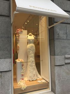 a wedding dress is displayed in a window with flowers on the outside and behind it are mannequins