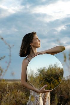 a woman holding a round mirror in her right hand and looking at the sky with clouds behind her