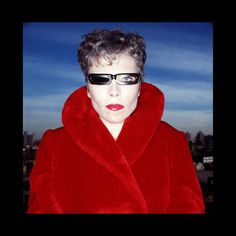 a woman wearing sunglasses and a red fur coat in front of a cityscape