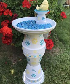 a white bird bath with blue and yellow flowers in the back ground next to red flowers