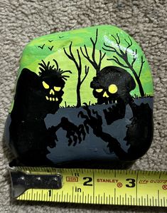 a black and green painted rock next to a measuring tape on the ground with two skulls in it