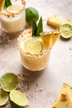 two glasses filled with drinks and garnished with limes, pineapple slices and coconut