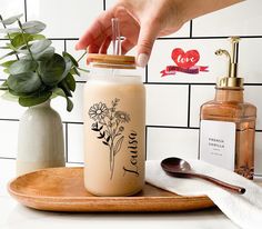 a person is holding a straw in a mason jar with flowers on it and a spoon next to the jar