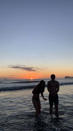 two people are standing in the water at sunset, one is wearing a bathing suit