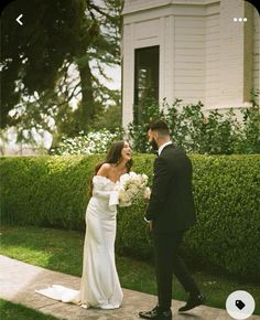 a bride and groom are walking together in front of a house with hedges on either side