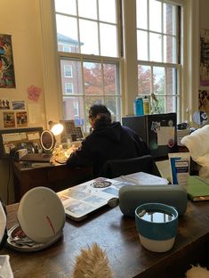 a person sitting at a desk in front of a window with lots of clutter on it