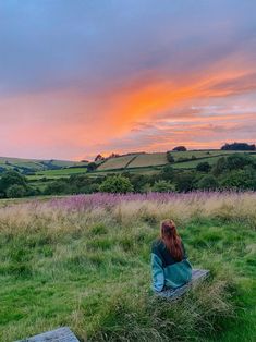 a woman sitting on top of a wooden bench in a lush green field under a colorful sky